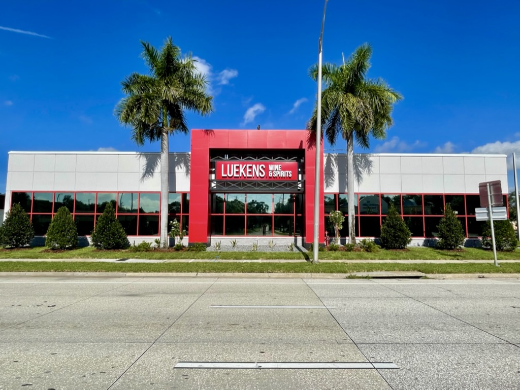 A retail store with a sign reading "Luekens Wine & Spirits," featuring a modern facade, red accents, and palm trees in front.