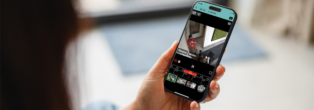 Persona que sostiene un teléfono inteligente que muestra una interfaz de aplicación de vigilancia, mostrando imágenes de una cámara de seguridad de una persona vestida de rojo y un perro cerca de una puerta.