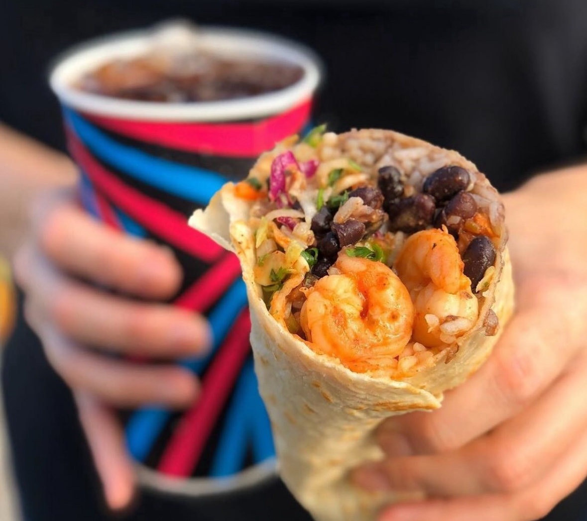 A person holds a shrimp and black bean burrito with colorful fillings in one hand and a cup of soda with a vibrant pattern in the other.