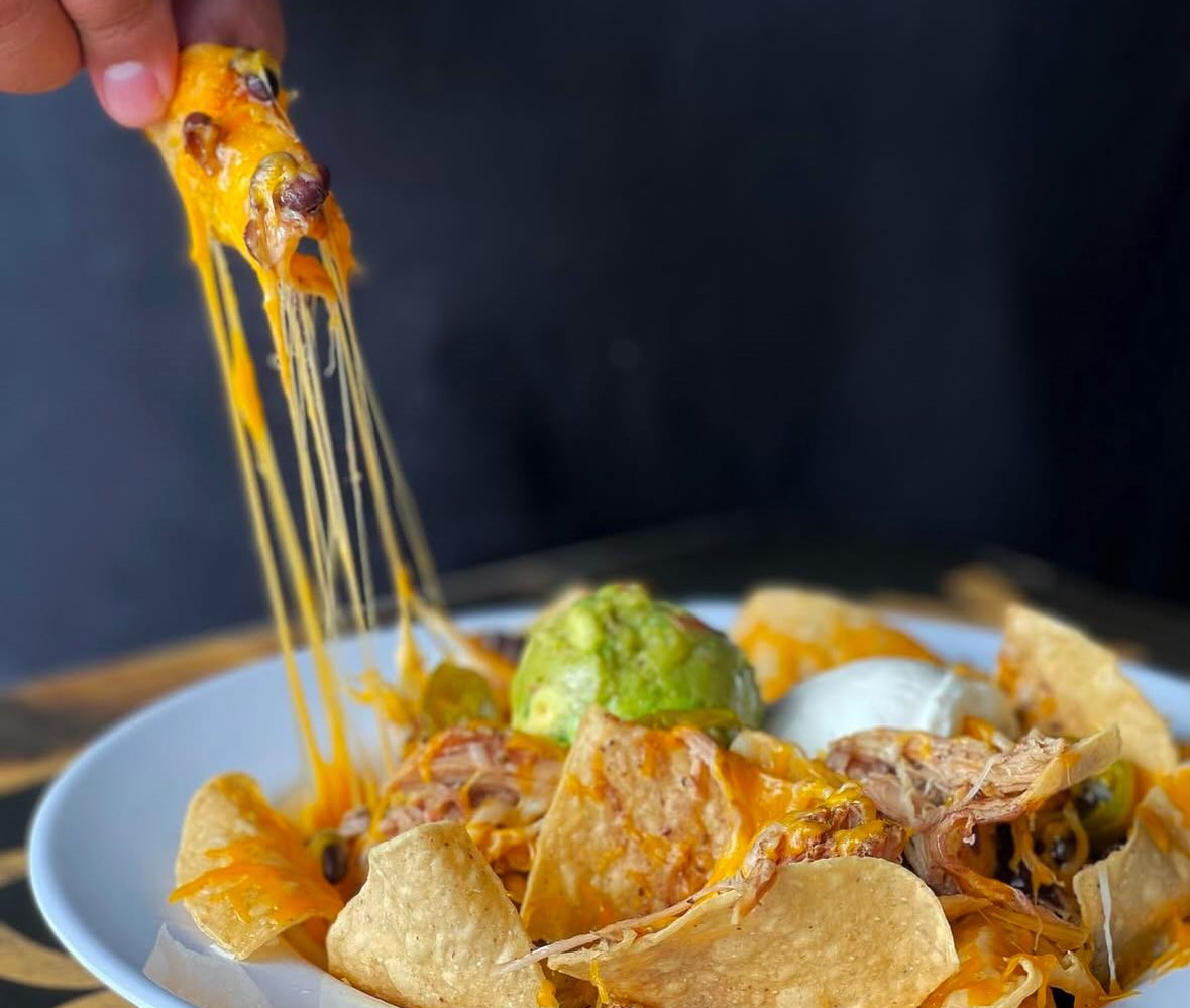 A plate of nachos with melted cheese, shredded meat, and guacamole. A hand lifts a nacho, stretching the cheese.