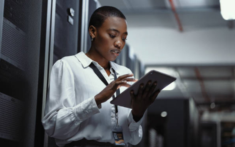 A person in a white shirt uses a tablet while standing in a server room with racks of equipment.