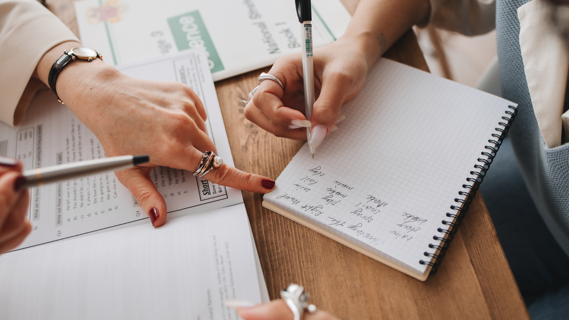 Three people are seated at a table with notebooks. One person writes with a pen in a spiral-bound notebook, while others point and discuss nearby open books. The scene suggests collaboration.