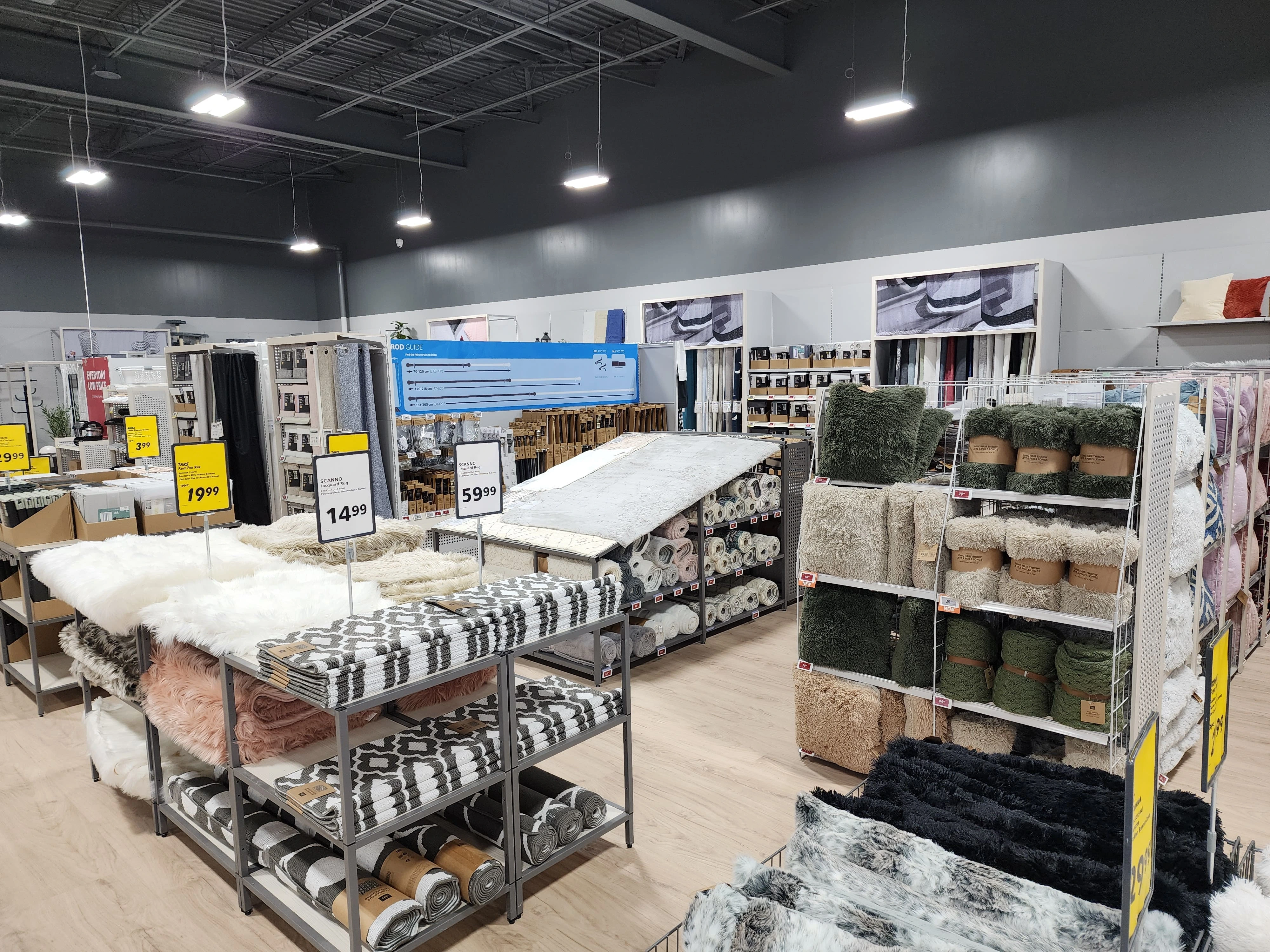 A store interior showing various rugs displayed on shelves and racks, with price tags visible. The space has high ceilings and overhead lighting.